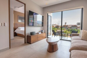 A modern hotel room with a bed, a TV, a wooden cabinet, a round coffee table, and a balcony with a view of the city. - Modern Versalles 2BR w/ Rooftop Pool & Views in Puerto Vallarta (Puerto Vallarta)