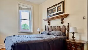 A bedroom with a bed, bedside tables, a window, and a wall-mounted shelf with decorative items. - Spacious 3BR /7 Guests/ Parking/Near NYC & Stadium (Bayonne)