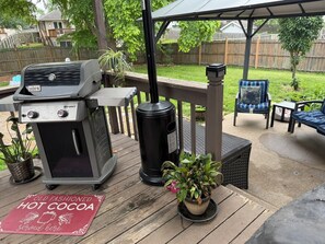 Um quintal com uma churrasqueira Weber Spirit, uma mesa de pátio, uma cadeira e uma placa com a inscrição 'Chocolate Quente Tradicional servido aqui'.