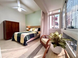 Ein Schlafzimmer mit einem Bett, einem Holzschrank, einem Sessel, einer Topfpflanze und einem Fenster mit Blick.
