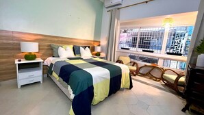 Quarto com cama, mesa de cabeceira, cadeira, janela com vista para prédios e ar condicionado.