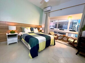Ein Schlafzimmer mit einem Bett, einem Nachttisch, einem Sessel, einem Fenster mit Blick auf Gebäude und einer Klimaanlage.