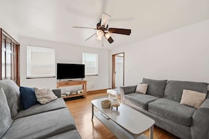 Ein modernes Wohnzimmer mit Flachbildfernseher, zwei grauen Sofas, einem Couchtisch und einem Deckenventilator.