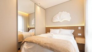 Quarto de hotel com um espelho grande, cama com lençóis brancos, cabeceira de madeira e um objeto decorativo fixado na parede.