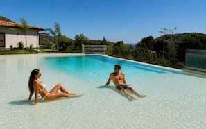 A swimming pool with two people sitting on the edge, surrounded by greenery and a house in the background. - Villa Heaven Nature (Lazio)