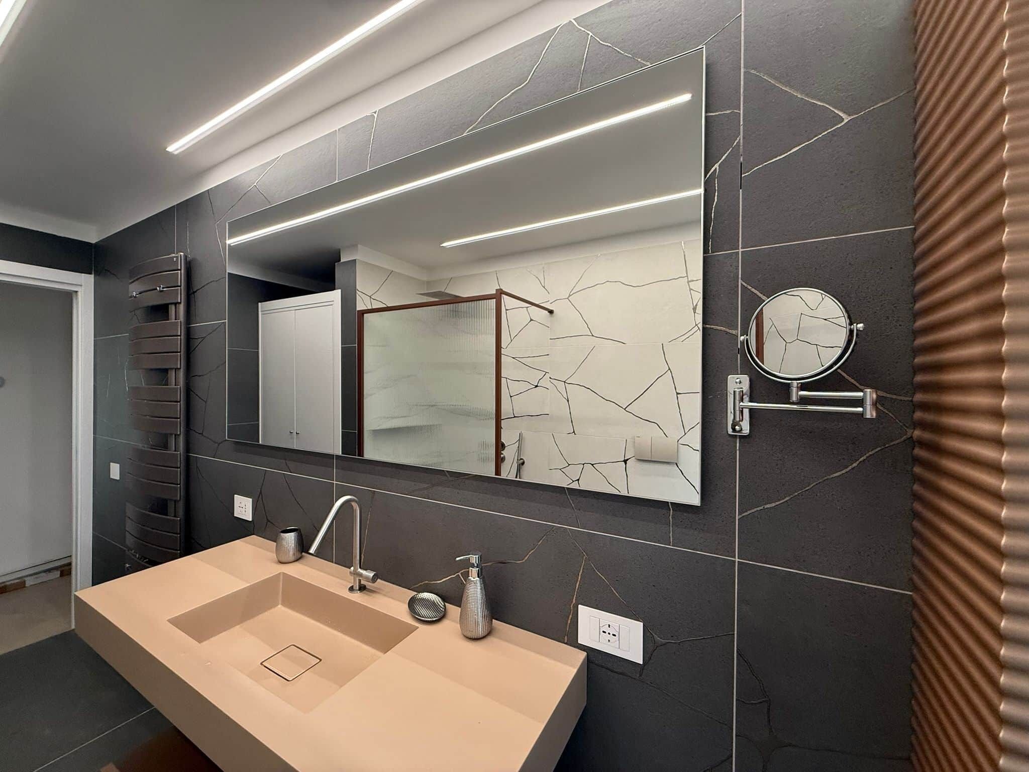A modern bathroom with a large mirror, a beige sink, and dark patterned tiles.