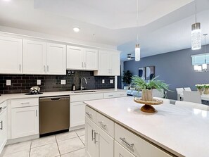 A modern kitchen with white cabinetry, a dishwasher, a black tiled backsplash, and a central island with a plant. - Storey Lake Cozy 3BR Condo, Lake View, By Disney (Kissimmee)