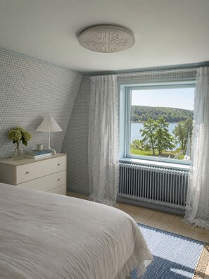 Ein Schlafzimmer mit einem großen Bett, einem Nachttisch mit Lampe, einer Kommode und einem Fenster mit Blick auf einen See und Bäume.