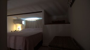 Ein Schlafzimmer mit Bett, Nachttisch, Lampe, einem Fenster mit Vorhängen und einem kleinen Regal.