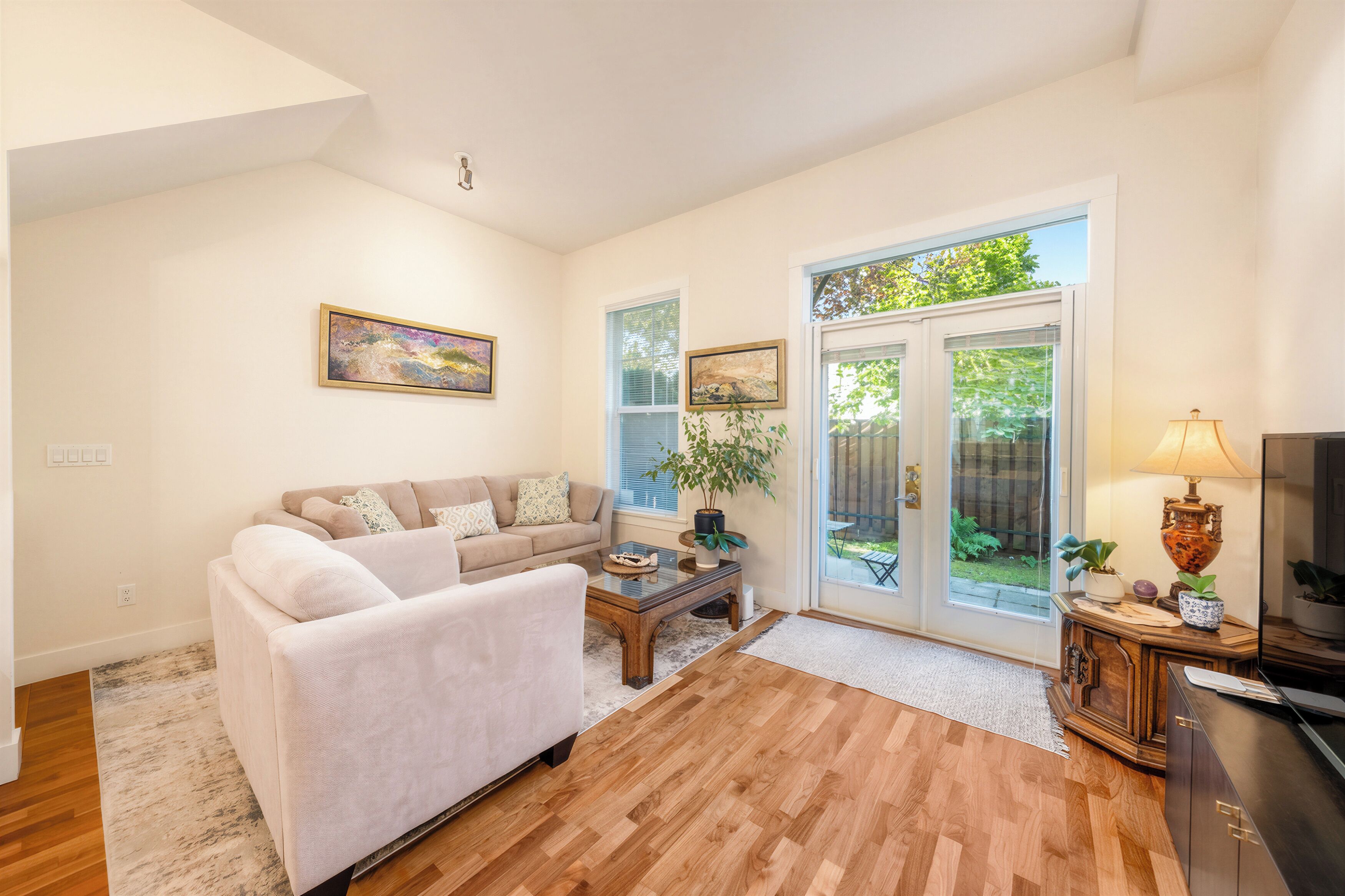 A living room with a sofa, coffee table, and a lamp. There is a sliding glass door leading to a garden.