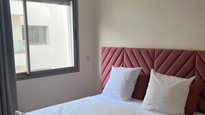 Ein Schlafzimmer mit großem Fenster, einem Bett mit weißer Bettwäsche und einem rosafarbenen Kopfteil sowie einer hängenden Lampenleuchte.
