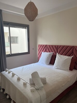 Ein Schlafzimmer mit großem Fenster, einem Bett mit weißer Bettwäsche und einem rosafarbenen Kopfteil sowie einer hängenden Lampenleuchte.