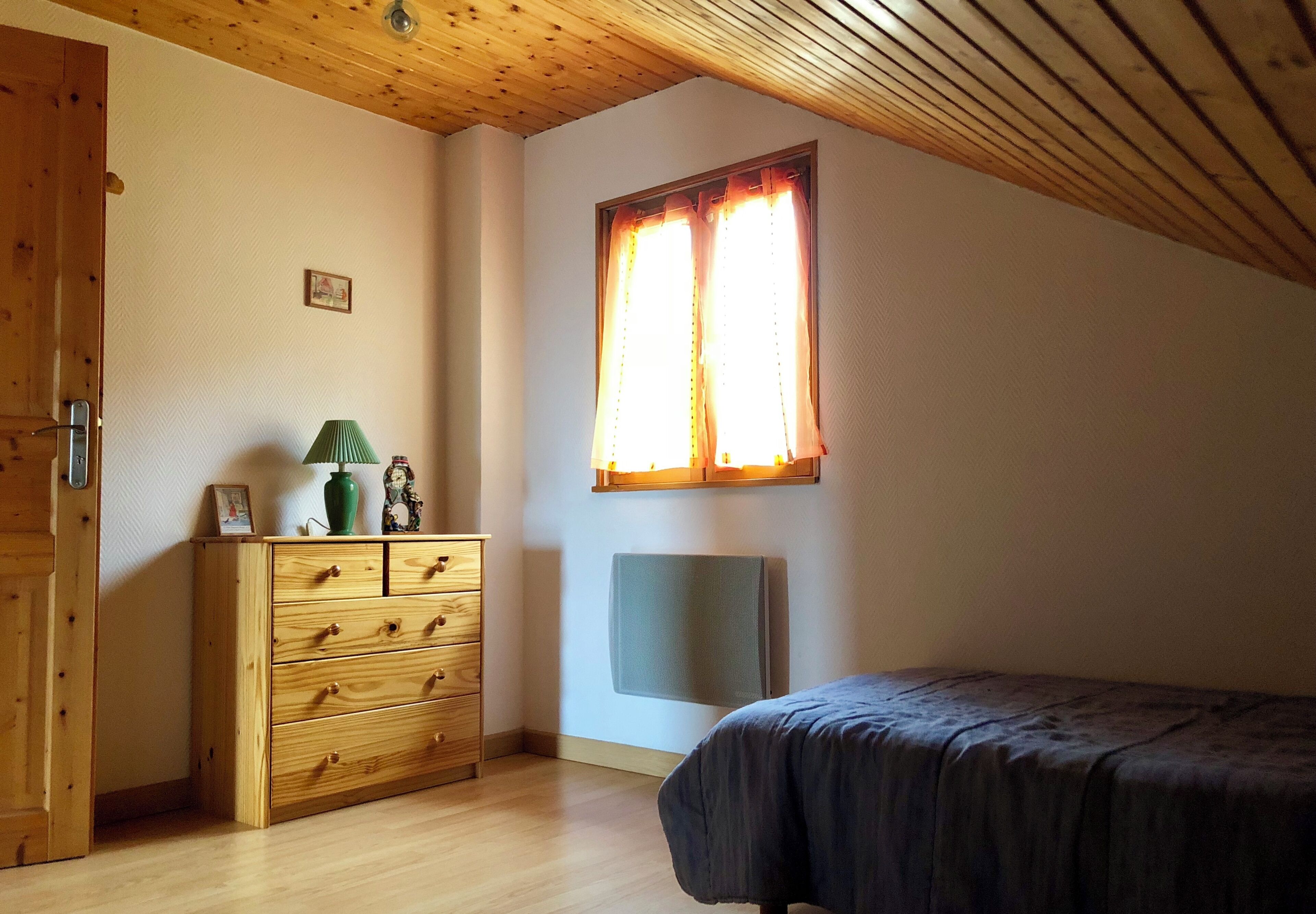 Camera da letto con soffitto in legno, un letto, una cassettiera, una finestra con tende e una TV a parete.