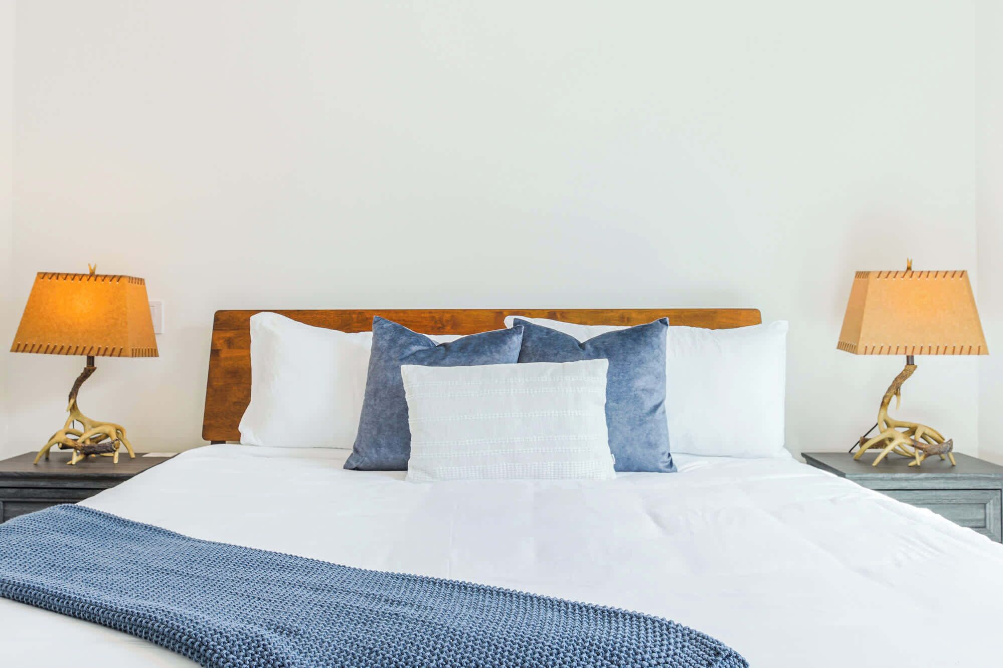 A neatly made bed with white linens, a blue knitted throw, and a wooden headboard. There are two bedside lamps with beige shades on either side of the bed.