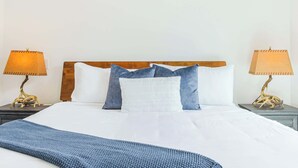 Cama bem arrumada com lençóis brancos, um cobertor azul de malha e cabeceira de madeira. Há duas luminárias laterais com abajures bege de cada lado da cama.