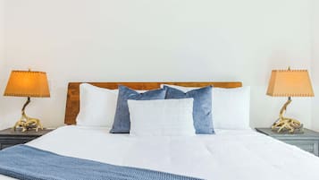 A neatly made bed with white linens, a blue knitted throw, and a wooden headboard. There are two bedside lamps with beige shades on either side of the bed.