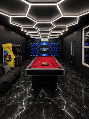Una mesa de billar con superficie de fieltro rojo, rodeada por una sala oscura con un diseño de techo hexagonal y un letrero que dice 'GAMEROOM'.