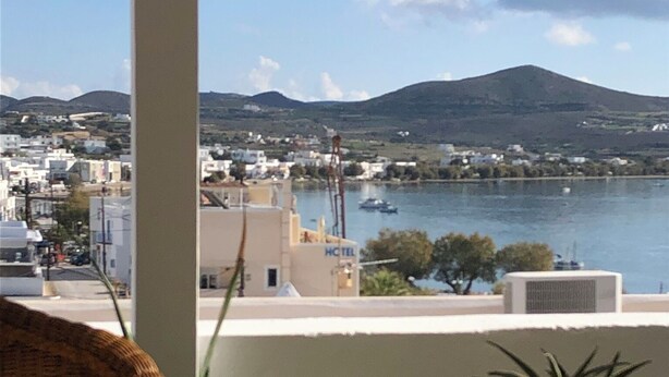 A balcony with potted plants, a wicker chair, and a view of a coastal town with buildings and a mountain in the distance. - Celia amazing view at the port of Adamas (Adamantas)