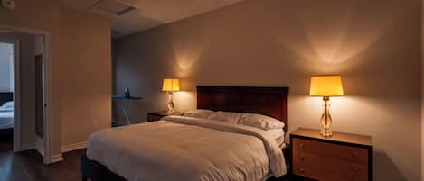 Une chambre à coucher avec un grand lit, deux tables de chevet avec des lampes, une tête de lit en bois et une porte donnant sur une autre pièce.
