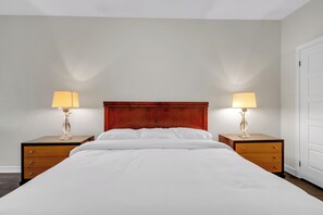 Een hotelkamer met een houten hoofdeinde, twee nachtkastjes met lampen, een bed met wit beddengoed en een witte deur.