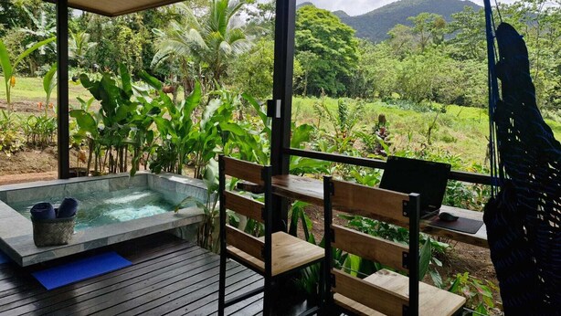 A wooden deck with a hot tub, wooden chairs, and a table overlooking a lush garden and mountains. - 1BR bungalow with plunge pool, outdoor shower & Arenal Volcano view (La Fortuna)