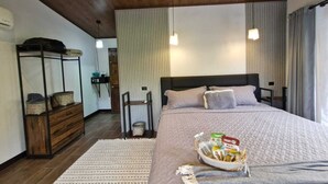 A bedroom with a wooden ceiling, a bed with a grey bedspread, a bedside table, and a nightstand. There is a small window with curtains, a wooden dresser, and a wall-mounted shelf with items on it. - 1BR bungalow with plunge pool, outdoor shower & Arenal Volcano view (La Fortuna)