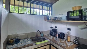 A kitchen with wooden cabinets, a microwave, a coffee maker, and a window with a view of greenery. - 1BR bungalow with plunge pool, outdoor shower & Arenal Volcano view (La Fortuna)