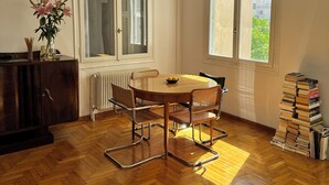 A dining area with a wooden table and chairs, a radiator, a window with a view, and a stack of books. - Quiet Designer Home, 2BR, Sunset Balcony • Kypseli (Athina)
