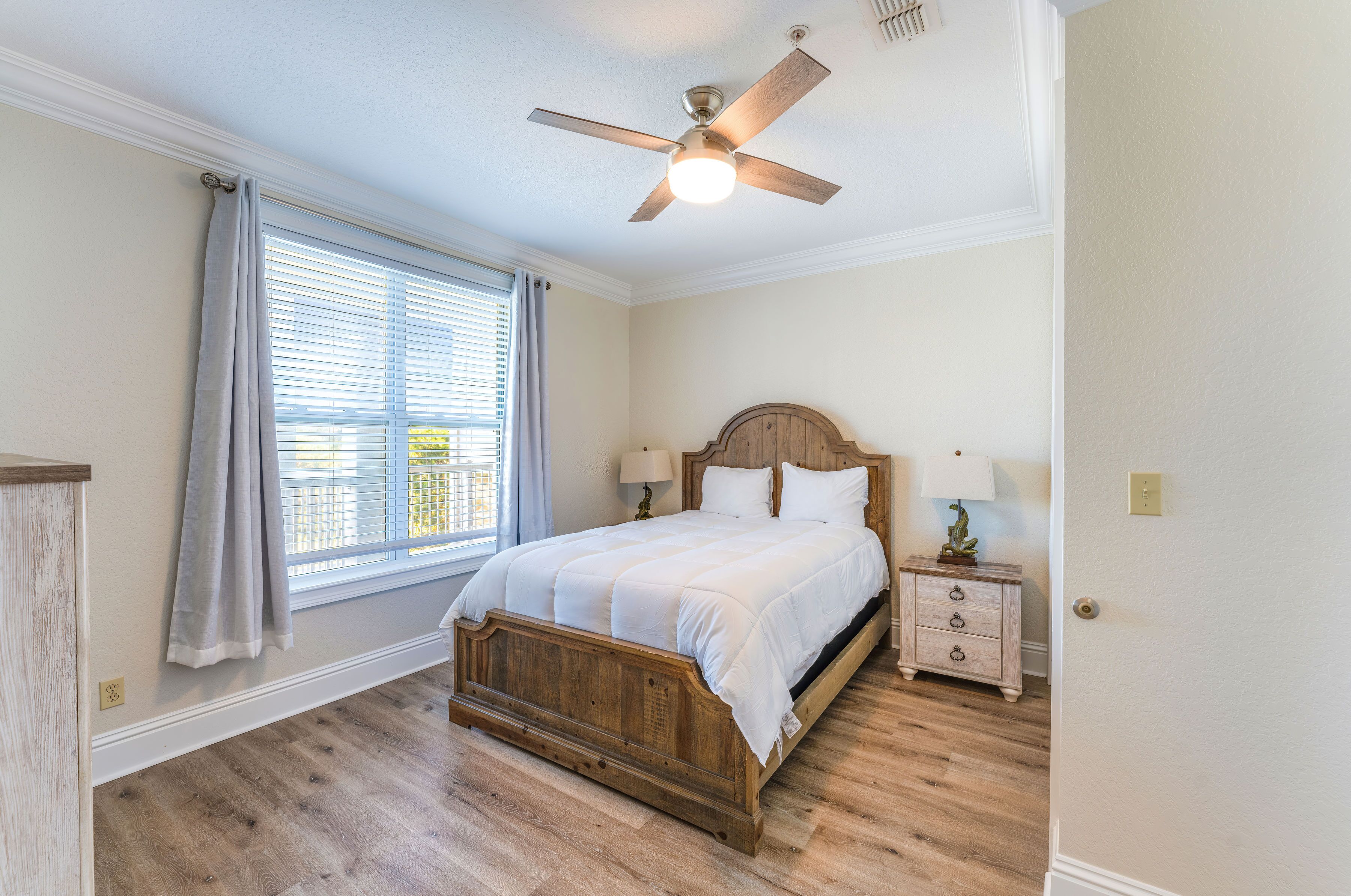 A bedroom with a wooden bed, a nightstand with a lamp, a ceiling fan, and a window with curtains.