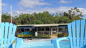 A swimming pool with blue chairs in the foreground, a building with a green roof in the background, and lush greenery surrounding the area. - The Dreamcatcher House (San Pedro)