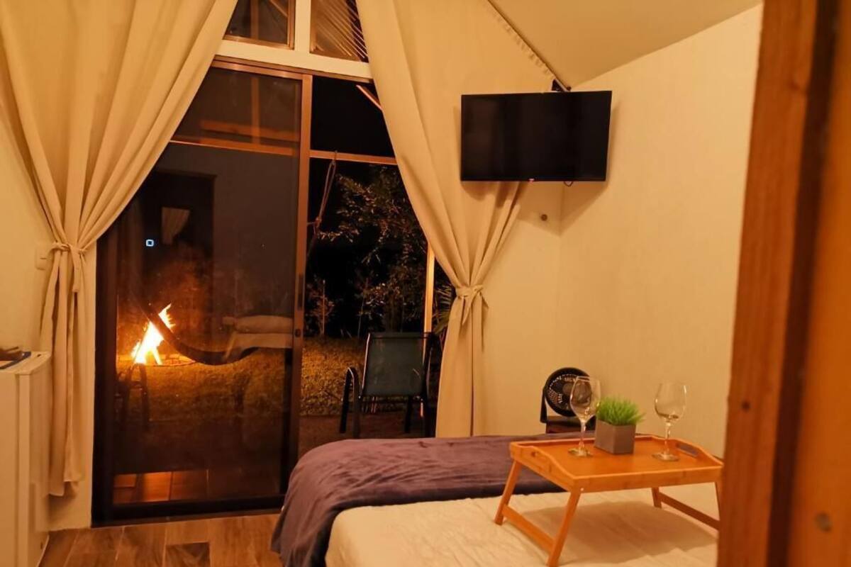 A bedroom with a bed, a TV mounted on the wall, a sliding glass door leading to an outdoor area with a fire, and a small table with a glass and a plant.