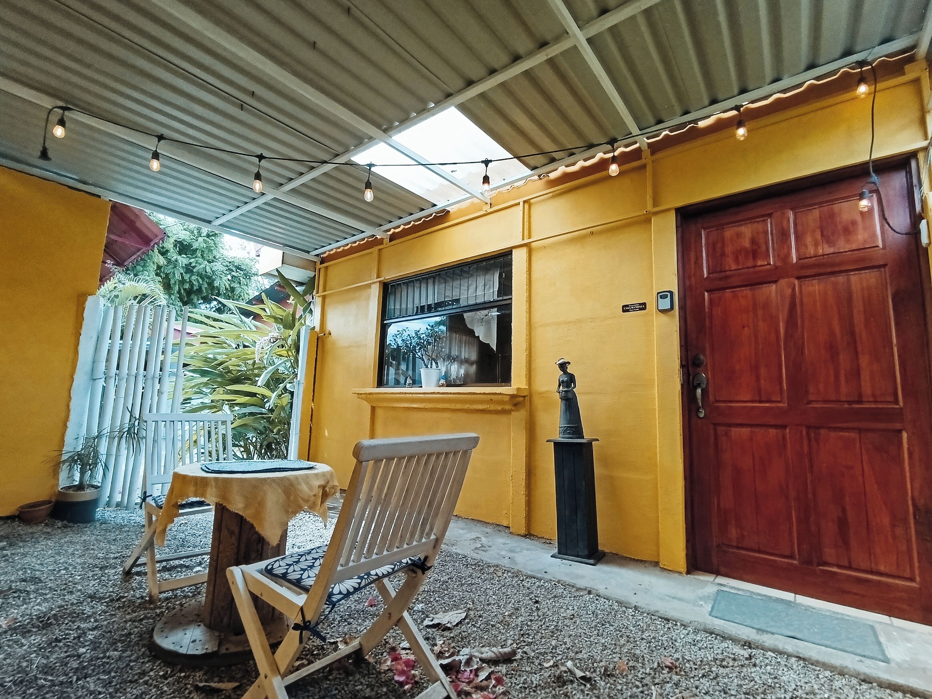 Un bâtiment jaune avec une porte en bois, une fenêtre et une statue. On y trouve une chaise et une table à l’extérieur.