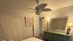 Et soveværelse med seng, ventilator, kommode og spejl.