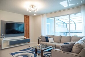 A modern living room with a flat-screen TV, a sofa, a coffee table, and a large window. - Spacious Townhouse w/ Pool & Free Parking Near Disney (Davenport)
