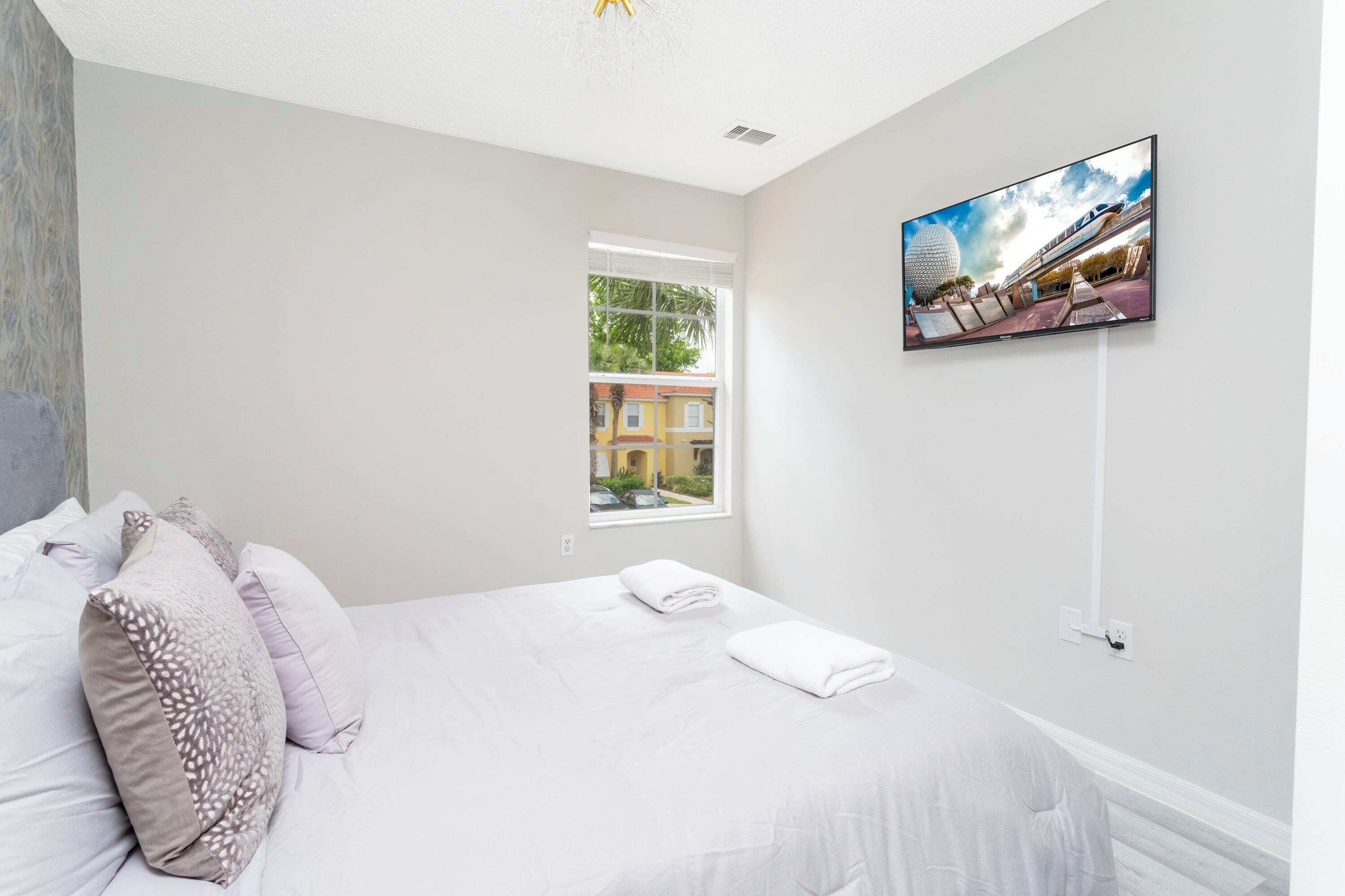 A bedroom with a bed, pillows, a TV on the wall, and a window with a view of buildings.