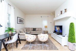 A living room with a sofa, two chairs, a TV, a plant, and a dining table.