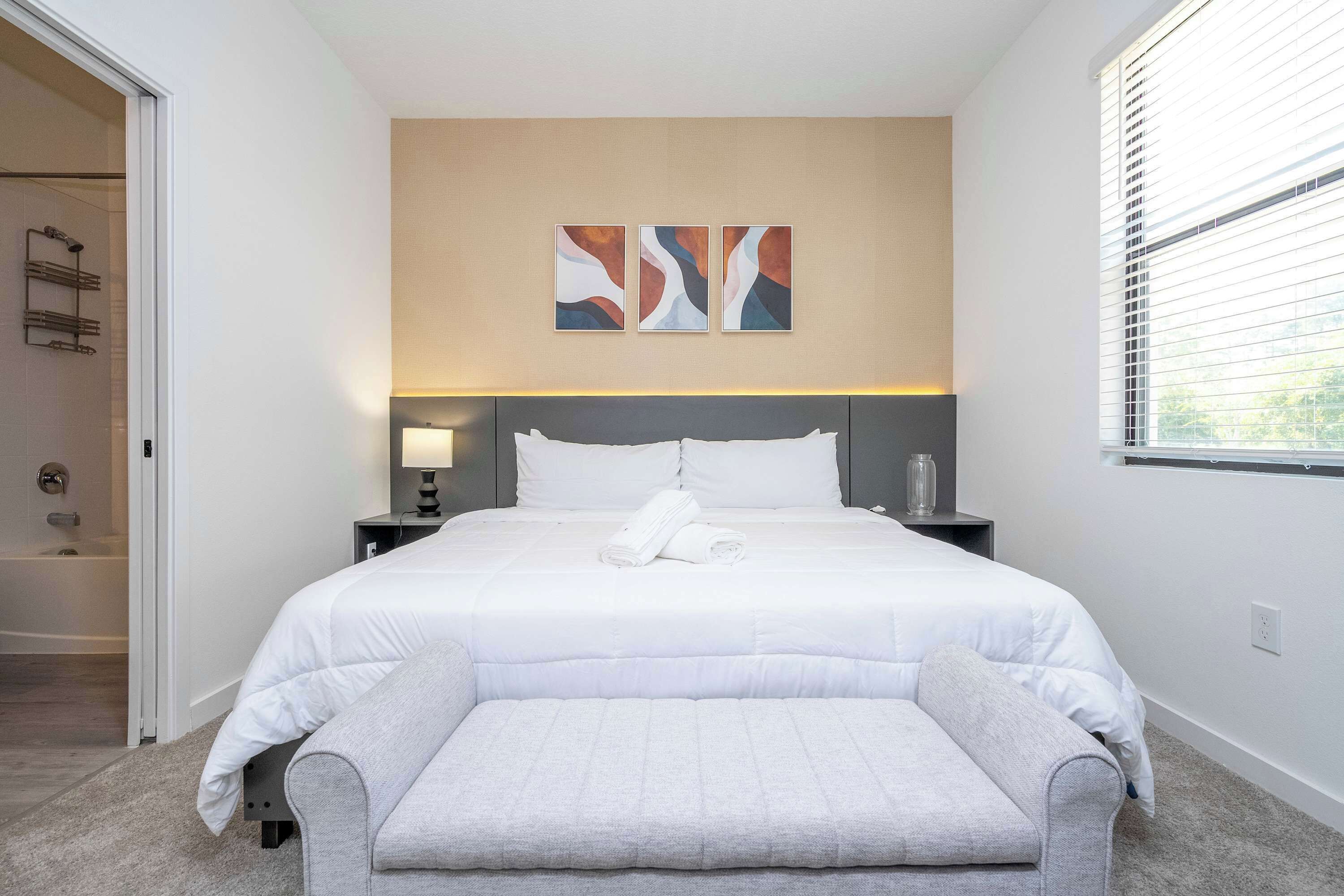 Une chambre d’hôtel moderne avec un grand lit, une table de chevet, un banc et une fenêtre avec des stores.