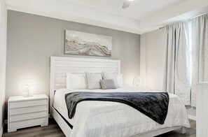 Ein Schlafzimmer mit einem weißen Bett, einem Nachttisch mit Lampe, einem Wandbild und einem Fenster mit Vorhängen.