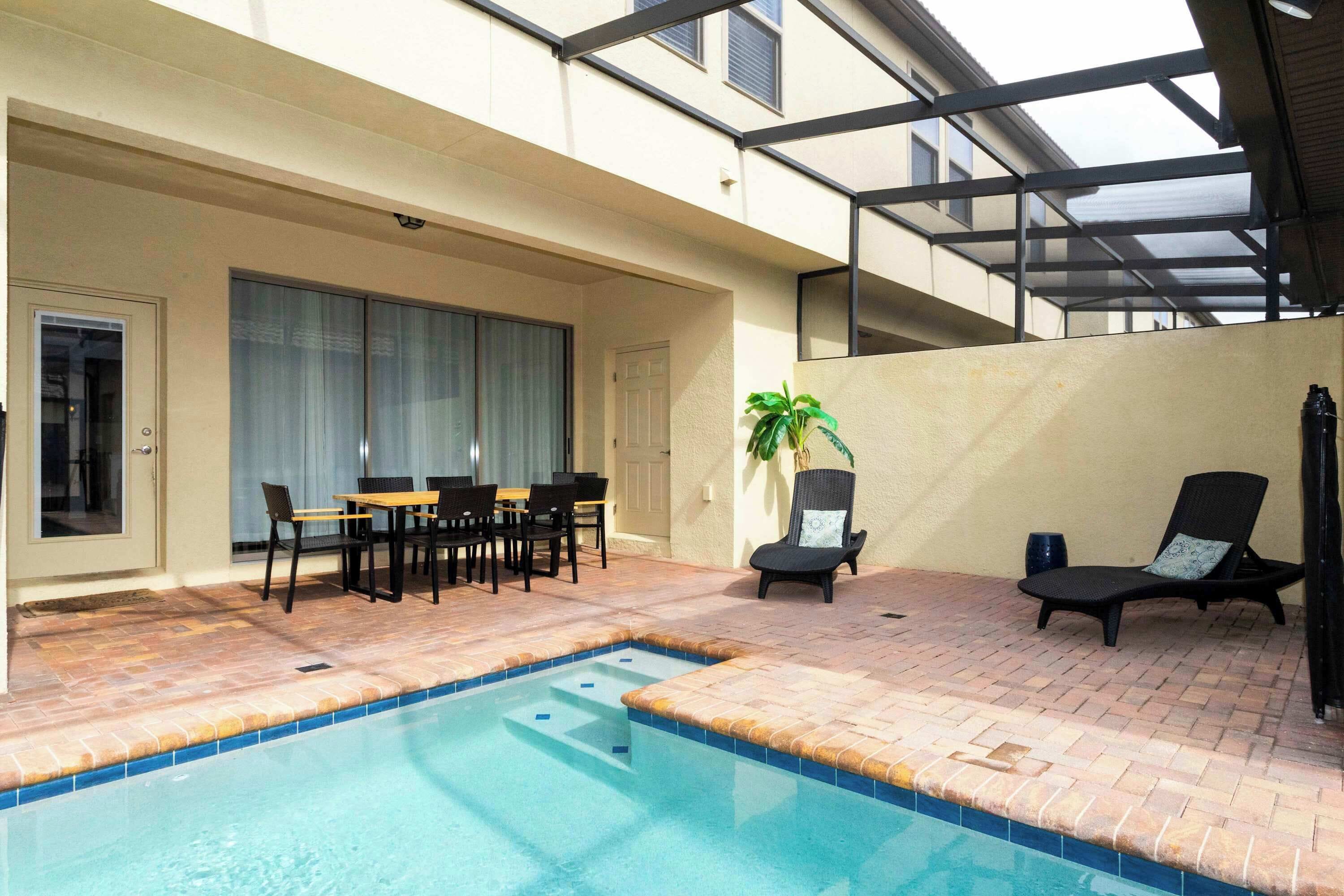 A swimming pool with a tiled edge, a covered patio area with a table and chairs, and a lounge chair.