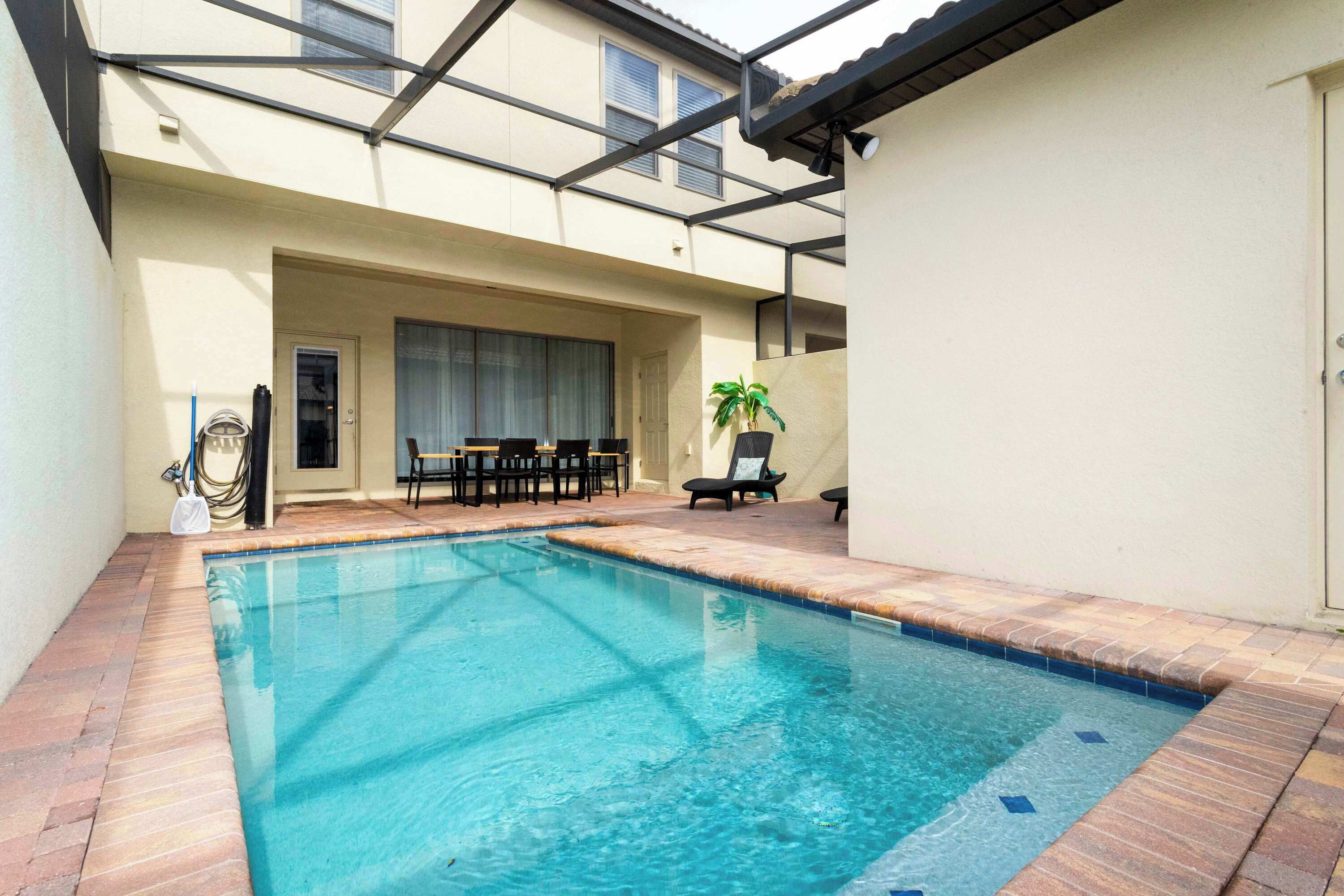 A swimming pool with a tiled edge, a covered patio area with outdoor furniture, and a potted plant.