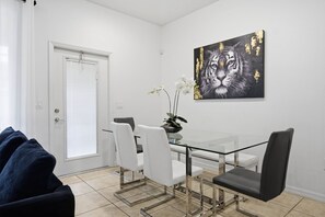 Un comedor con mesa y sillas de vidrio, una orquídea en maceta y un cuadro con tema de tigre en la pared.