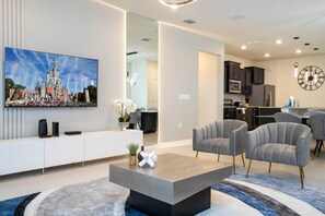 Ein modernes Wohnzimmer mit Flachbildfernseher, grauen Sofas, einem Couchtisch und einer Küche im Hintergrund.