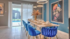 Zona pranzo con un tavolo lungo apparecchiato per sei persone, sedie in velluto blu, un vaso con fiori secchi e un grande quadro incorniciato appeso al muro.