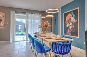 A dining area with a long table set for six, blue velvet chairs, a vase with dried flowers, and a large framed painting on the wall. - Family-friendly House With Private Pool & Game Room (Kissimmee)