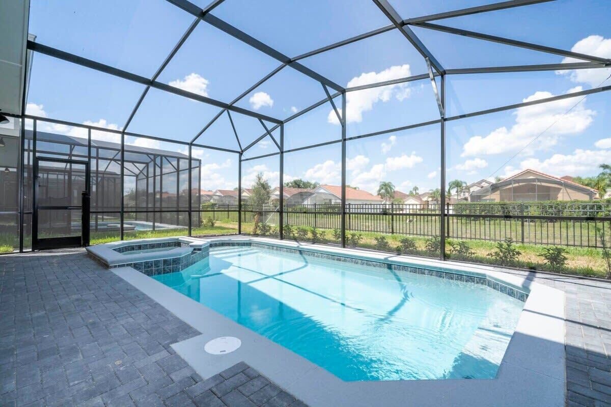A swimming pool with a glass enclosure, surrounded by a paved deck and a metal fence.