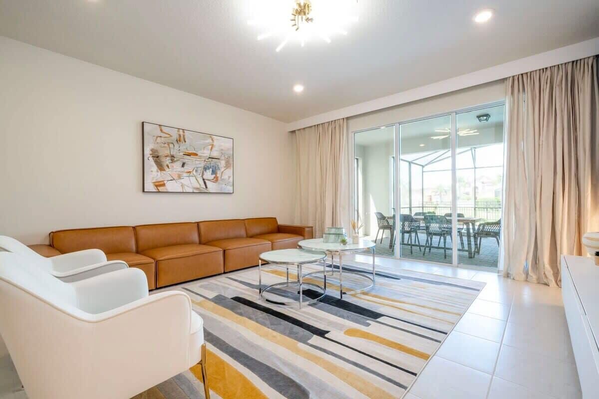A modern living room with a brown sofa, white armchairs, a glass dining table, and a large window with curtains.