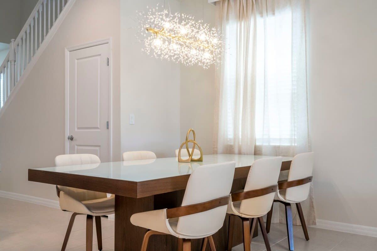 Een eetgedeelte met een houten tafel, witte stoelen, een kroonluchter en een trap op de achtergrond.