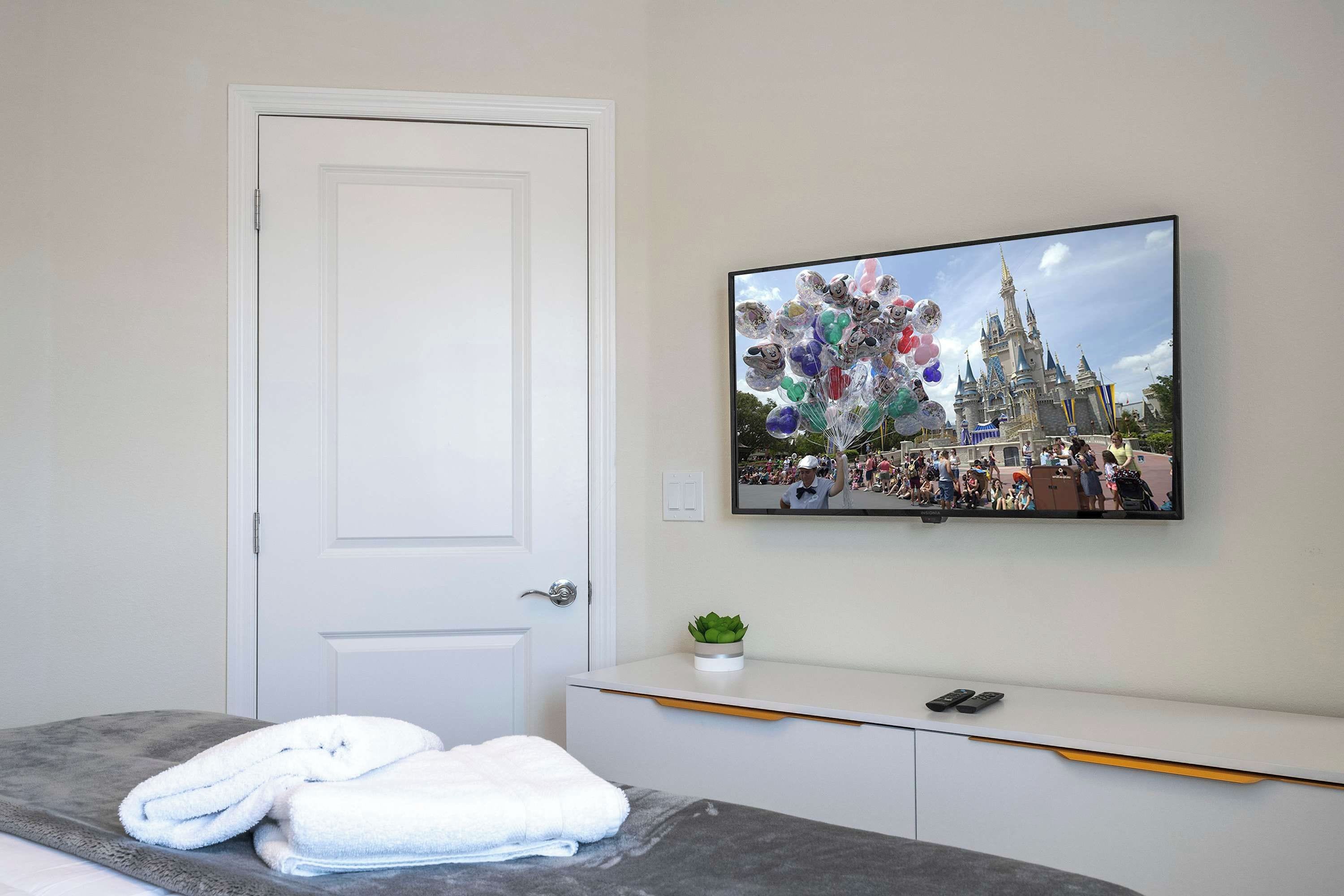 A hotel room with a white door, a flat-screen TV mounted on the wall, a grey sofa, and a small potted plant.