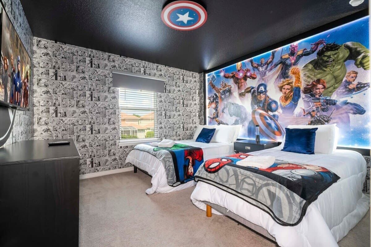Un dormitorio con dos camas, un mural grande con temática de superhéroes, una ventana con persianas y un escritorio.