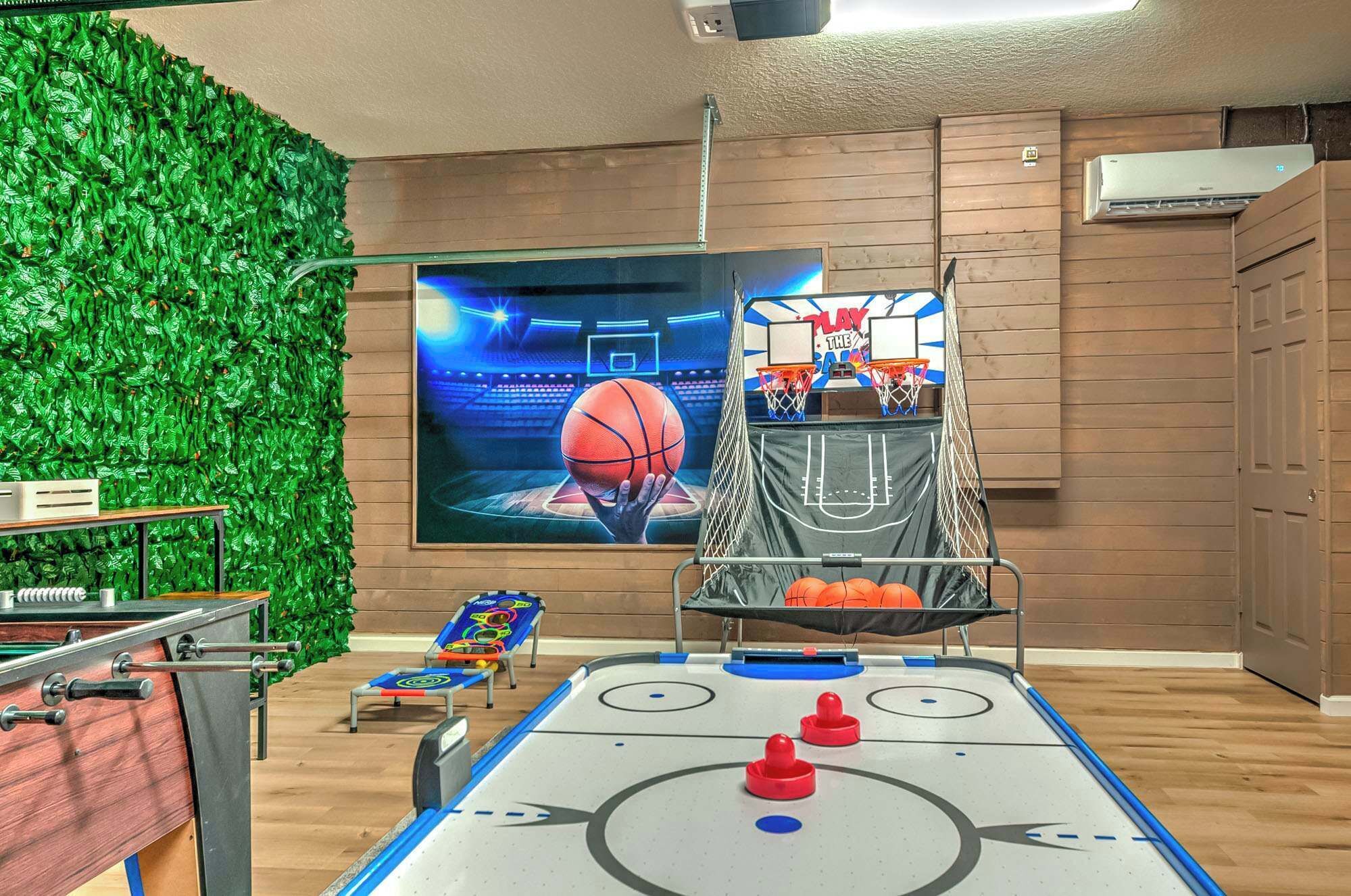 A room with a foosball table, a pool table, a green wall, and a basketball hoop.